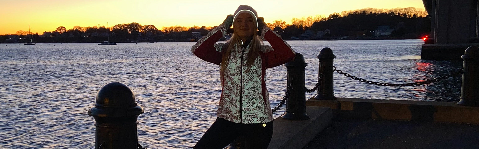 A woman standing on a pier at twilight. She is wearing a reflective running jacket and a reflective knit cap.