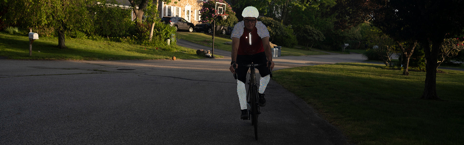 A man riding a bicycle towards the camera. He is wearing a reflective bike jersey, helmet cover, and leggings.