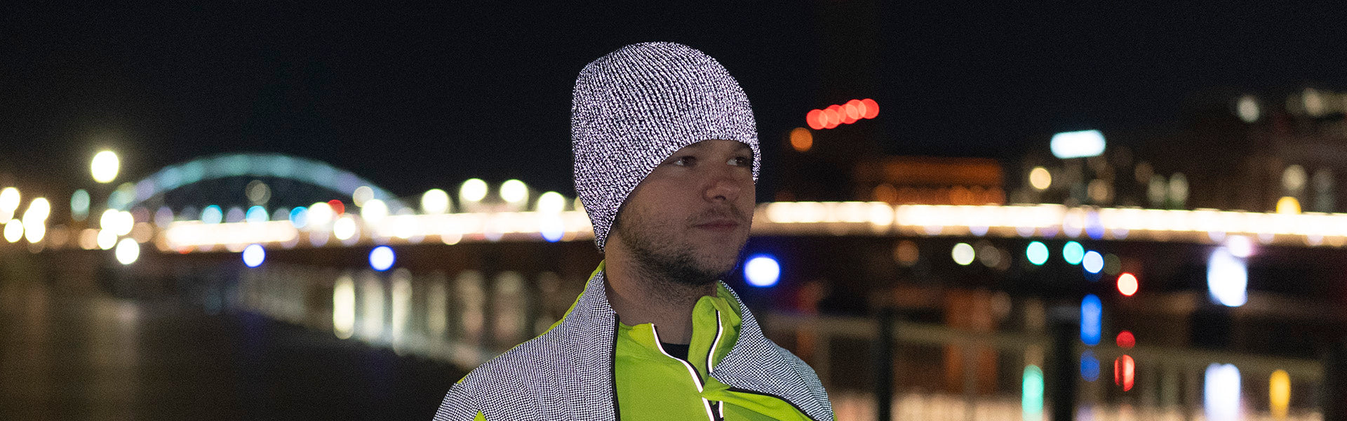 A man at night with a river and city skyline in the background. He is wearing a reflective beanie and a reflective softshell running jacket.