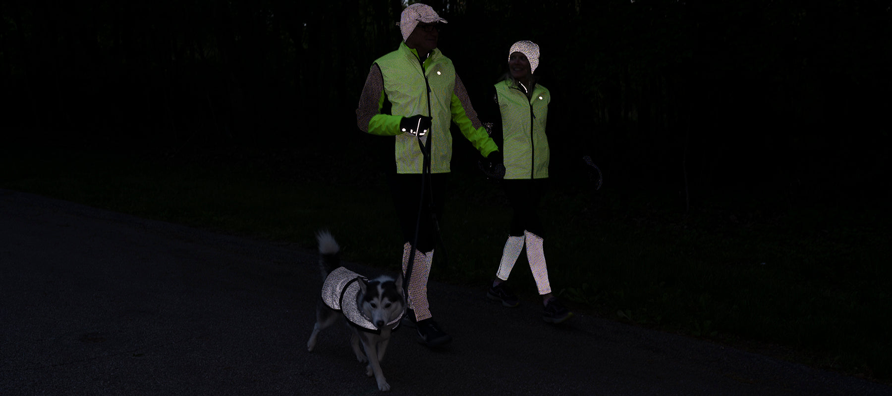 A man and a woman walking a dog at night. They are wearing reflective vests, leggings, and hats. The dog has a reflective jacket.