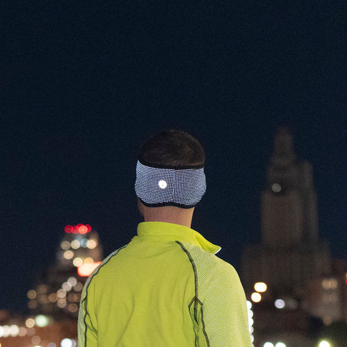 A man wearing a reflective headband at night. We see him from behind as he is gazing at an urban skyline in the distance.