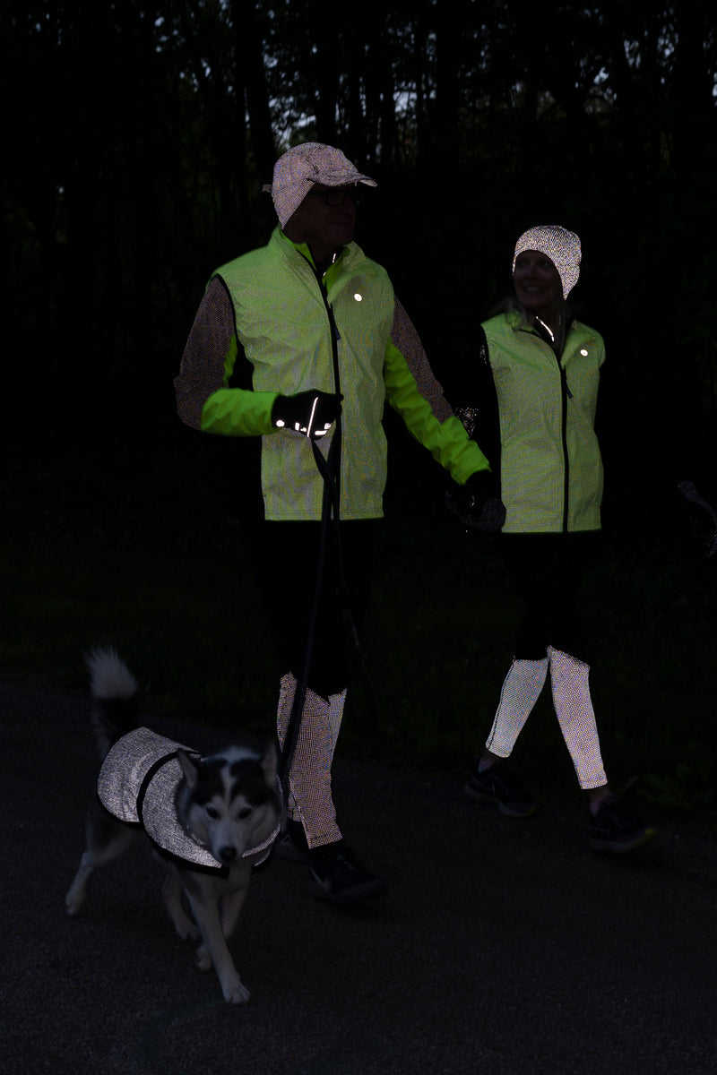 A man and a woman walking a dog at night. They are wearing reflective vests, leggings, and hats. The dog is wearing a reflective jacket.